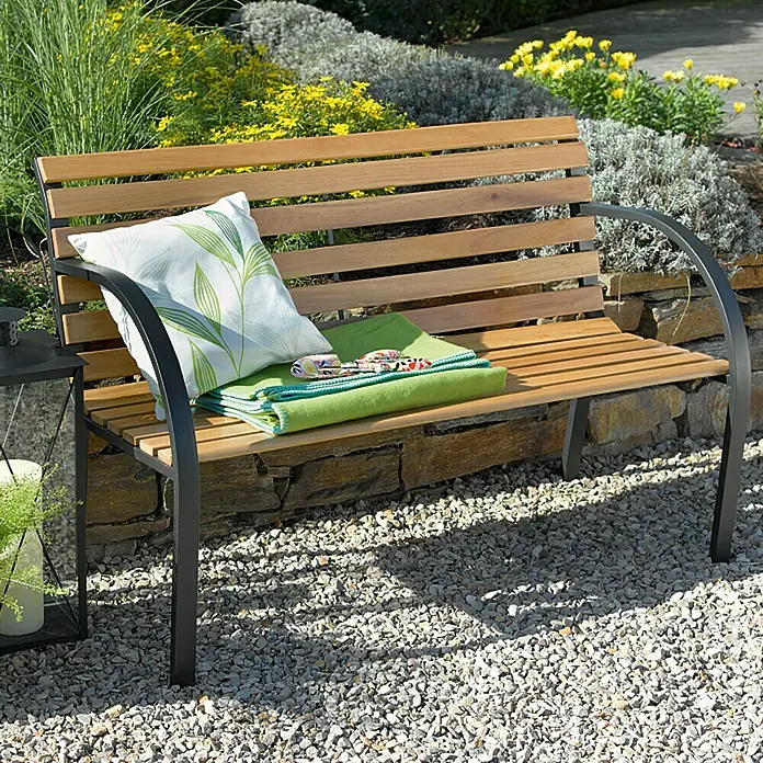 Sunfun Banco de jardín Saale - An x Pr x Al: 122 x 60 x 80 cm, Madera dura - Banco de jardín de madera y metal con almohada y manta en un camino de grava frente a macizos de flores.