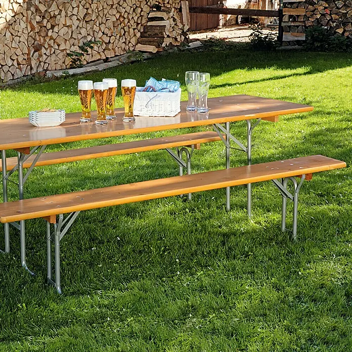 Sunfun Bierzeltgarnitur - 3 -tlg., Nadelholz/Stahl, Klappbar, Naturbraun/Silber, Tisch (L x B x H): 220 x 70 x 77 cm, Bank (B x T x H): 220 x 25 x 49 cm - Ein langer Biertisch mit Bänken steht im Grünen, Gläser und Snacks bereit für eine gesellige Runde im Freien.
