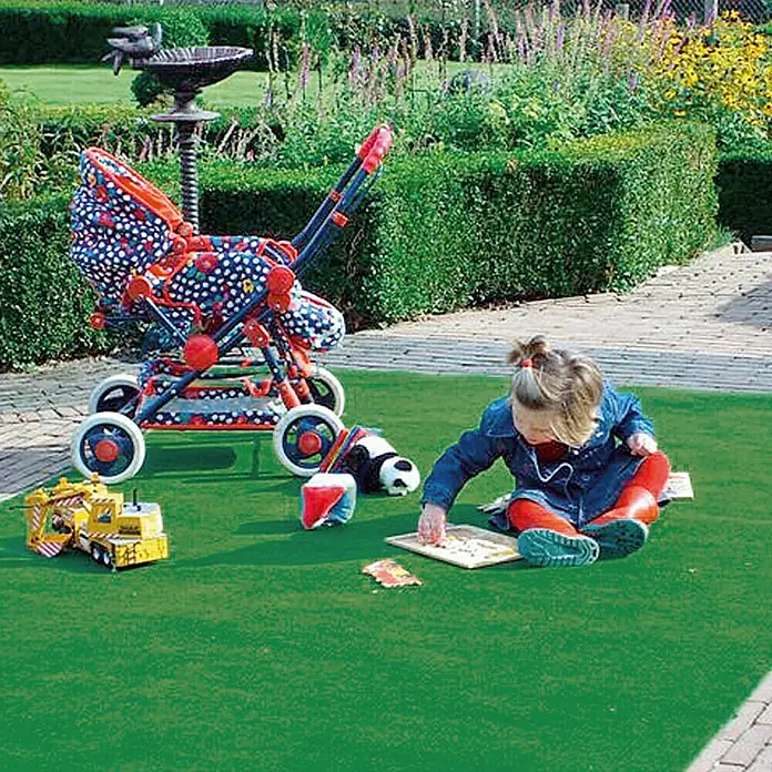 Rasenteppich Meterware Green - Breite: 150 cm, Grün, Mit Drainagenoppen - Ein farbenfroher Kinderwagen steht neben einem Mädchen, das auf einer grünen Fläche sitzt und spielt, was die Fantasie anregt.
