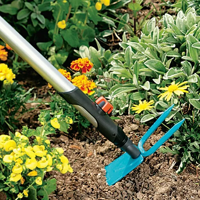 Gardena Combisystem Gartenhacke - Arbeitsbreite: 7,5 cm, Blattform: Gerade - Nahaufnahme eines blauen Gartenwerkzeugs mit zwei Zinken und einem Schaber, das in Blumenerde neben bunten Blumen gesteckt wird.