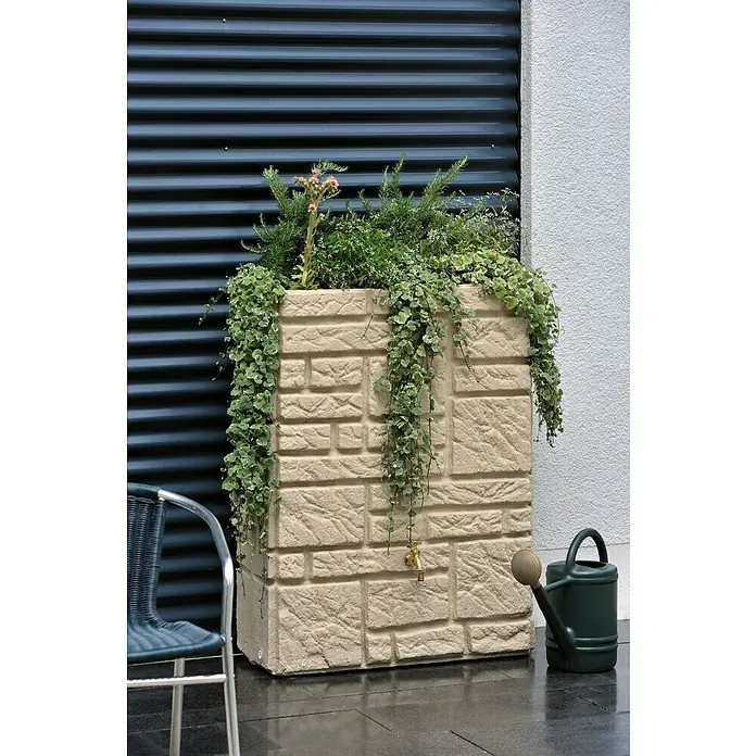 Arves Regenspeicher Maurano - 300 l, Sandsteinfarben, 1 Stk. - Ein quadratischer Pflanzkübel in Steinoptik mit üppigen Grünpflanzen, zusammen mit einer Gießkanne, ziert eine Terrasse.
