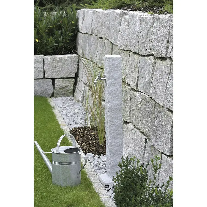 Arves Wasserzapfstelle Natura - Granitfarben, Höhe: 98 cm - Nahaufnahme einer Wasserstelle aus Granit mit Wasserhahn, umgeben von Grünpflanzen und einem Rasen. Eine Gießkanne steht daneben.