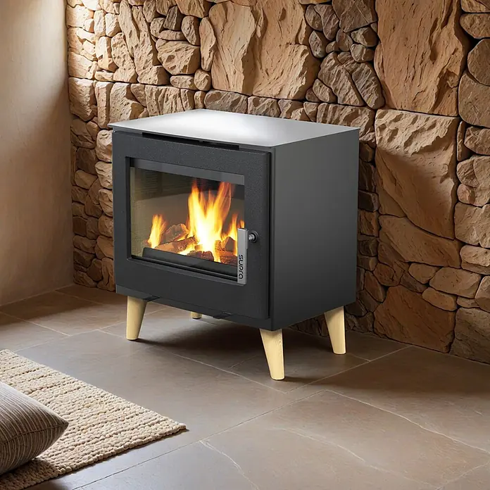 Supra Estufa de leña Emile - 6,1 kW, Volumen de calefacción: 250 m³, Negro - Estufa de leña gris oscuro con puerta de cristal, fuego visible y patas de madera clara, en una habitación con pared de piedra rústica.