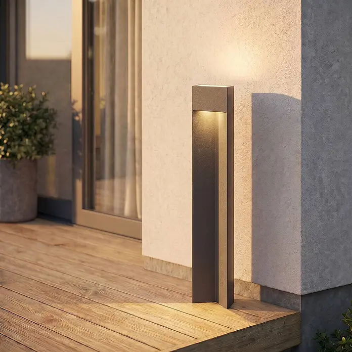 Lucande Außenlaterne Kaholo -  - Dunkelgraue rechteckige Wegeleuchte, die eine Holzterrasse und angrenzende Wand mit warmem Licht beleuchtet.