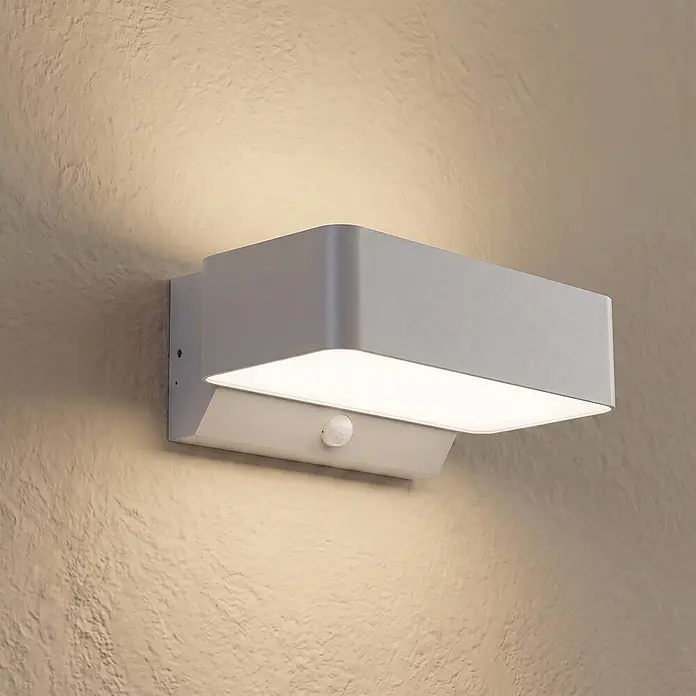Lindby LED Wandleuchte aussen in silber matt, weiß, mit Bewegungsmelder, -  - Graue, rechteckige Wandleuchte mit Bewegungsmelder, die eine strukturierte Wand beleuchtet.