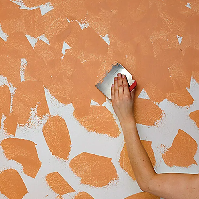 Erfurt Vliestapete Variovlies Flat Classic - Weiß, Uni, 25 x 0,75 m - Eine Hand mit Spachtel trägt orangefarbene Farbe auf eine weiße Wand auf.