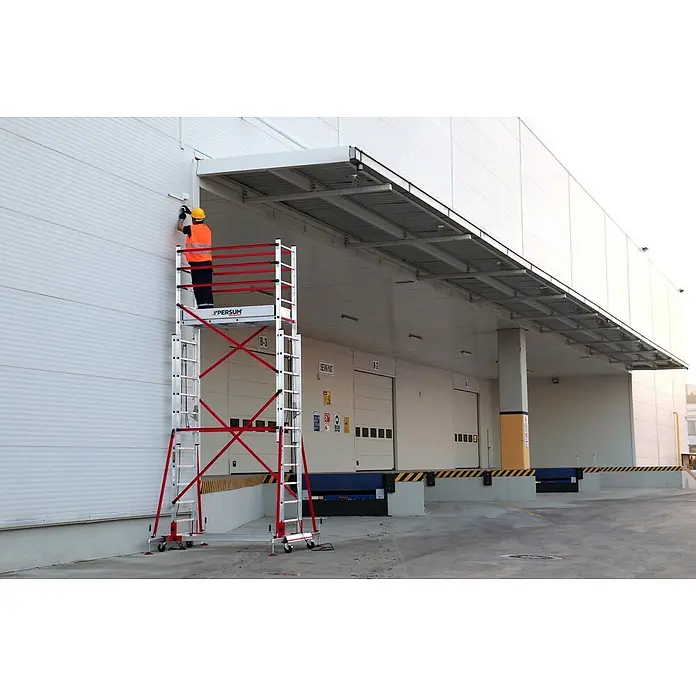 Andamio plegable Teletower - Altura de trabajo: 4,75 m, Ancho de la plataforma: 1,55 m, Longitud de la plataforma: 0,6 m - Andamio móvil plateado y rojo con un trabajador en su plataforma, utilizado para el mantenimiento de una pared de edificio.