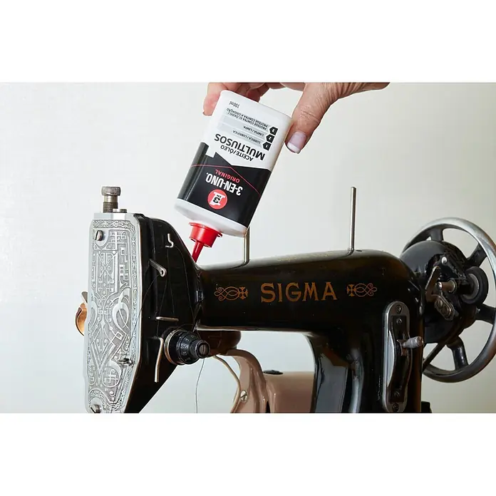 3 en 1 Lubricante - 100 ml, Apto para: Herrajes - Una mano aplica aceite multiusos 3-EN-UNO de una botella blanca con boquilla roja en una máquina de coser Sigma vintage negra.