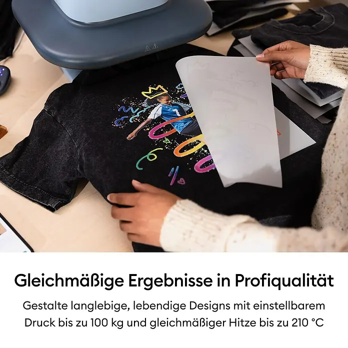 Cricut Transferpresse Autopress 2 - 53,5 x 41,3 x 29,4 cm, Drucktechnologie: Wärmeübertragung, Slate Blue - Eine Person legt ein farbenfrohes Transferdesign auf ein schwarzes T-Shirt, daneben eine Heißpresse zur Übertragung des Designs.