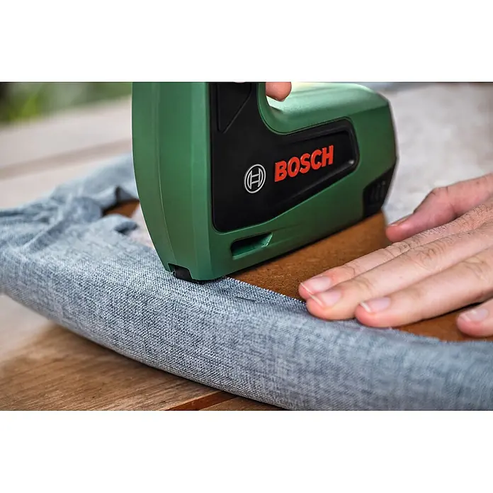 Bosch Akku-Tacker Tweo - 4 V, 1 Akku, 2 Ah, Klammertiefe: 4 mm - 10 mm - Grünes und schwarzes Bosch-Tacker von einer Hand verwendet, um grauen Stoff auf braune Oberfläche zu heften.