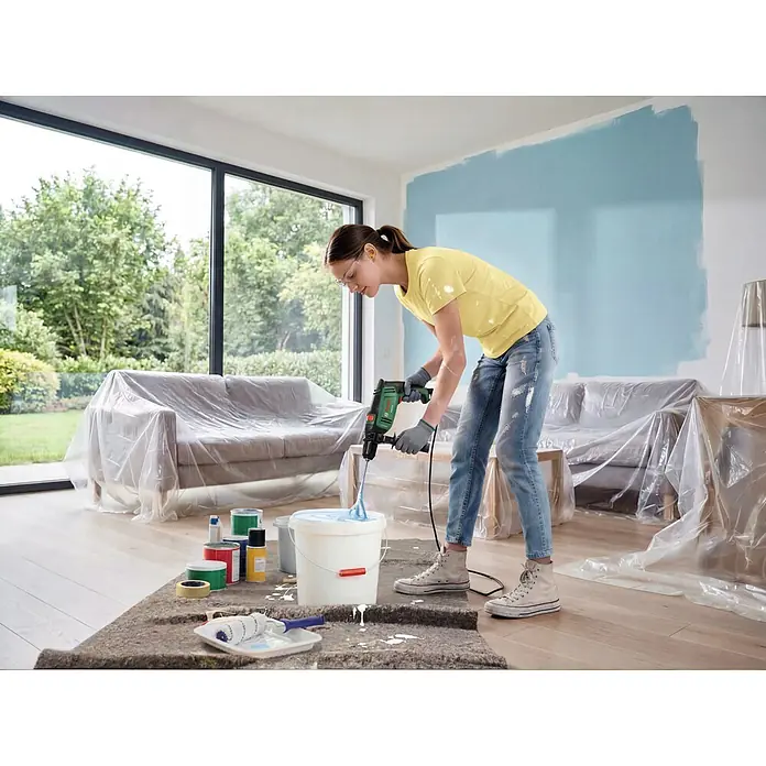 Bosch Taladro percutor PSB750 - 550 W - 750 W, Portabrocas de sujeción rápida - Una mujer usa un taladro verde y negro con mezclador para remover pintura azul claro en un cubo, preparándose para pintar una pared.