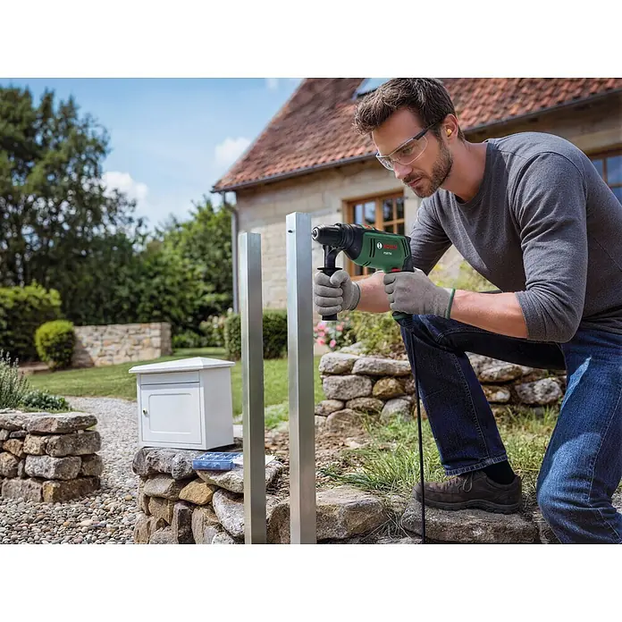 Bosch Taladro percutor PSB750 - 550 W - 750 W, Portabrocas de sujeción rápida - Un hombre con gafas de seguridad y guantes utiliza un taladro Bosch PSB 750 verde y negro para perforar un poste metálico al aire libre.