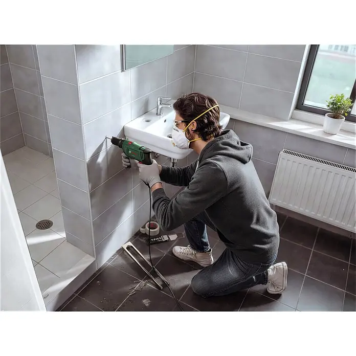 Bosch Taladro percutor PSB650 - 400 W - 650 W, Portabrocas de sujeción rápida - Una persona en un baño utiliza un taladro Bosch verde y negro para hacer un agujero en una pared de azulejos grises.