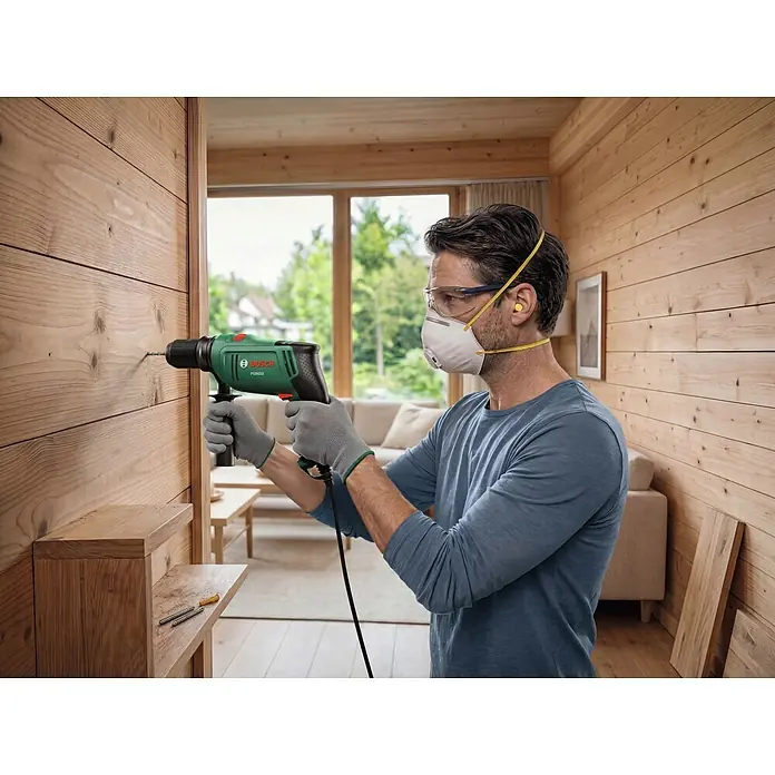 Bosch Taladro percutor PSB650 - 400 W - 650 W, Portabrocas de sujeción rápida - Un hombre con gafas de seguridad y mascarilla usa un taladro percutor Bosch PSB 860 verde para perforar una pared de madera.