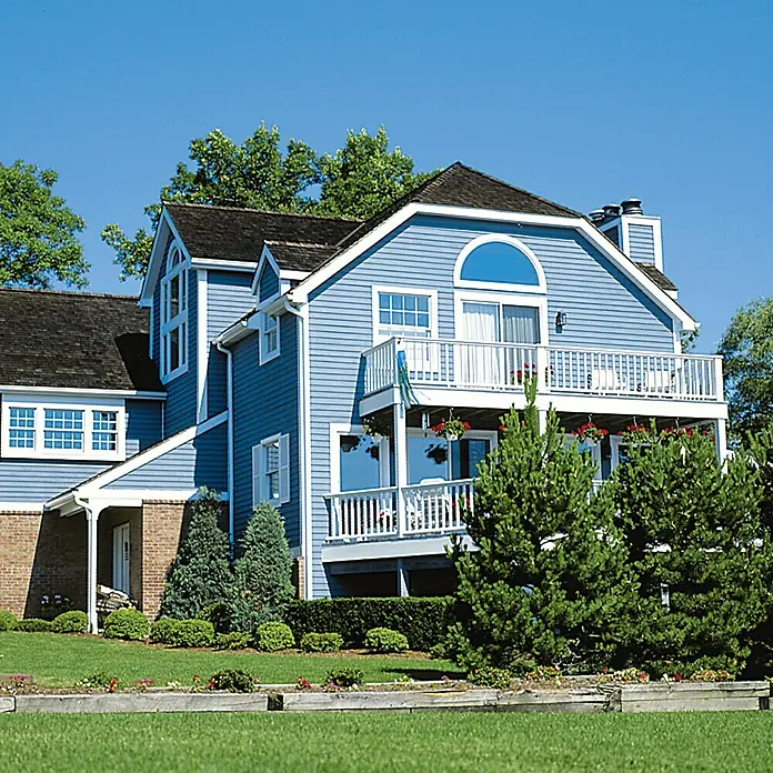 swingcolor Mix Holzschutzfarbe zum Mischen lassen - Basismischfarbe 4, 2,5 l, Seidenglänzend - Blaues Haus mit zwei Balkonen, umgeben von üppigem Grün und einem strahlend blauen Himmel, der die Architektur betont.