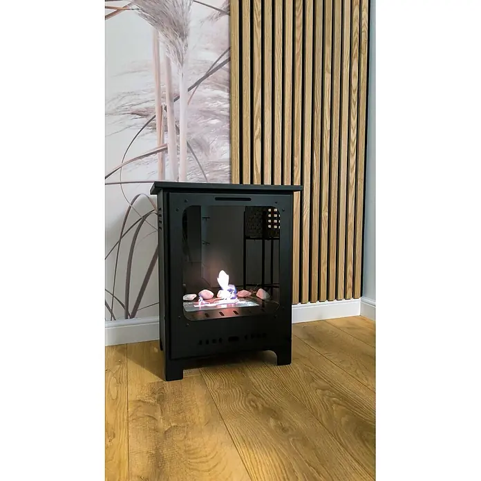 Bysil Kaminofen - Material Verkleidung: Stahl, Schwarz - Ein schwarzer Bio-Kamin mit kleiner Flamme und Dekorsteinen, auf Holzboden vor einer gemusterten Wand.