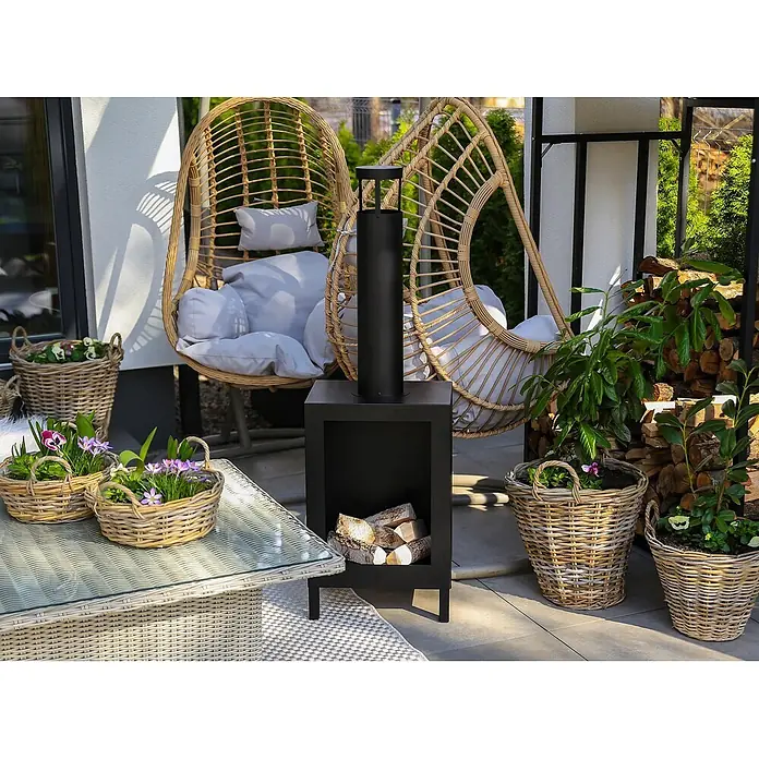 Bysil Kaminofen - Material Verkleidung: Stahl, Schwarz - Schwarze Outdoor-Feuerstelle mit Kamin und Holzablage, auf einer Terrasse zwischen zwei Rattan-Hängesesseln.