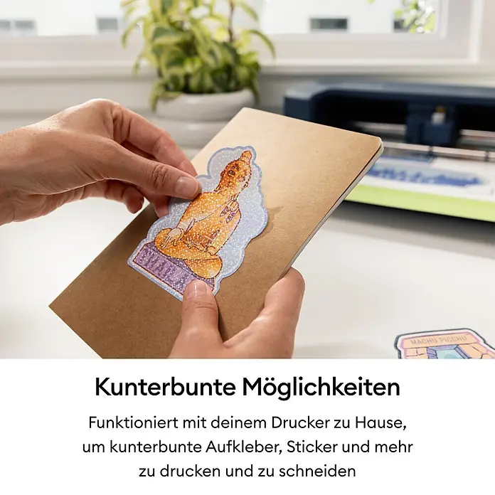Cricut Maker Schneideplotter Maker 5 Essentials-Bundle - Schiefer, Bluetooth, Schnittbreite: 30,5 cm, Schnittlänge: 3,6 m - Person bringt einen holografischen Glitzer-Buddha-Aufkleber auf ein Notizbuch an. Eine Schneidemaschine ist sichtbar, was die Erstellung individueller Aufkleber zeigt.