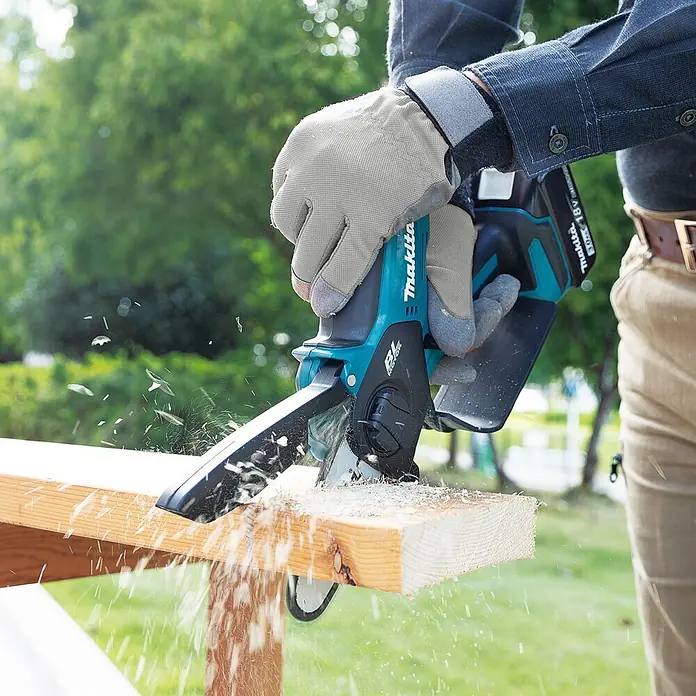 Makita LXT 18V Einhand-Akku-Kettensäge DUC101RX06 - 18 V, Li-Ionen, 2 Akkus, Schwertlänge: 10 cm - Eine Person mit Handschuhen sägt mit einer blau-schwarzen Makita-Mini-Kettensäge ein Holzbrett, wobei Späne entstehen.