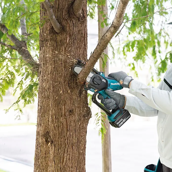 Makita LXT 18V Einhand-Akku-Kettensäge DUC101RX06 - 18 V, Li-Ionen, 2 Akkus, Schwertlänge: 10 cm - Person in Handschuhen schneidet mit einer türkis-schwarzen Makita Kettensäge einen Ast ab, Späne fliegen.