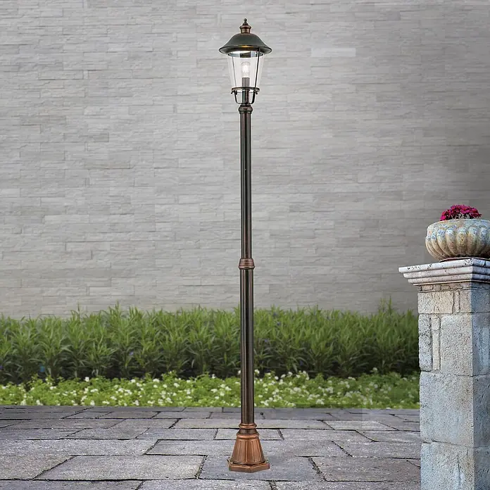 Orion Leuchten Außenlaterne Kenzo - 60 W, Farbe Gehäuse: Kupfer, 25 x 25 x 216 cm, Ohne Leuchtmittel - Klassische bronzene Außenlaterne mit beleuchteter Glühbirne, auf einem gepflasterten Weg in einer Gartenumgebung.