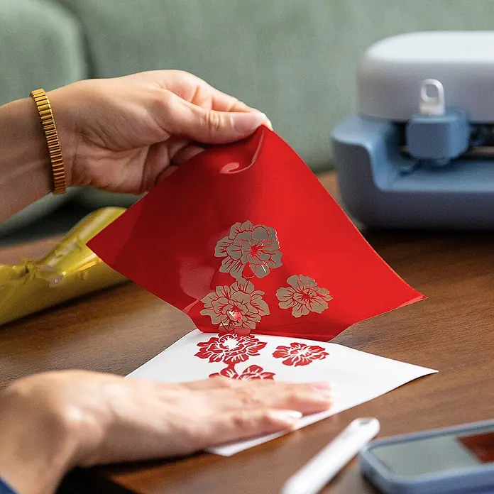 Cricut Joy Ersatzklinge Premiumfeinschnittklinge - Passend für: Cricut Joy™, Cricut Joy™ 2 und Cricut Joy Xtra™ - Hände ziehen rote Transferfolie mit silbernen Blumendesigns von einem weißen Blatt ab, rote Designs werden sichtbar. Bastelprojekt.