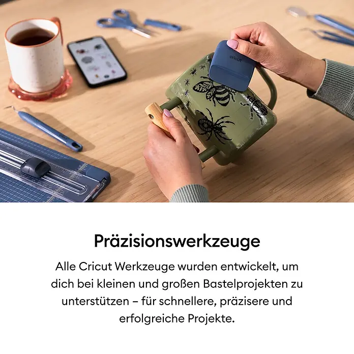 Cricut Plotter-Werkzeug Entgitterhaken - Geeignet für: Aufkleber, Blau - Eine Person nutzt ein blaues Cricut Schaberwerkzeug, um ein Design auf eine grüne Tasse aufzubringen, umgeben von Bastelwerkzeugen und einem Telefon auf einem Holztisch.