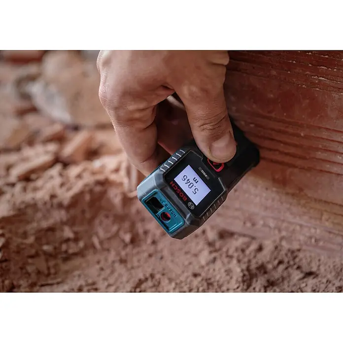 Bosch Professional Medidor de distancia láser GLM 50-21 - Gama de medición: 0,15 m - 50 m - Una mano sostiene un medidor láser Bosch negro y turquesa, que muestra '5,046 m', midiendo contra una superficie marrón rojiza.