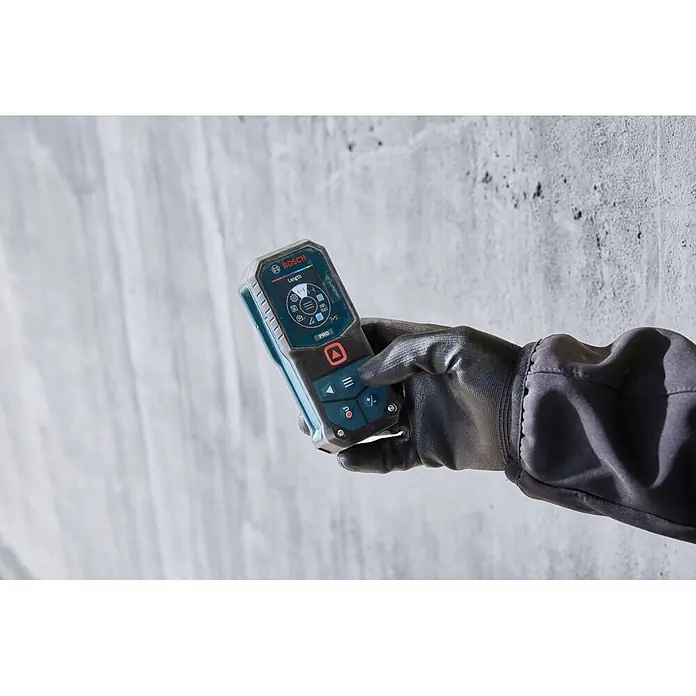 Bosch Professional Medidor de distancia láser GLM70-32 - Gama de medición: 0,05 m - 70 m - Una mano enguantada sostiene un medidor láser Bosch negro y azul con pantalla digital, listo para usar contra una pared texturizada.
