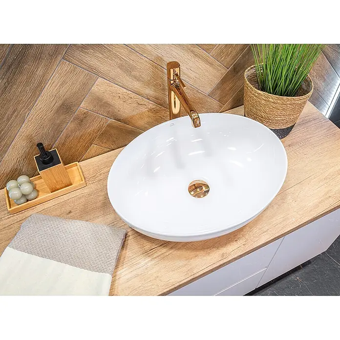 Bad Land Waschtischarmatur Pola hoch, Rosegold, mit Click-Clack -  - Modernes weißes Keramik-Aufsatzwaschbecken mit goldenem Wasserhahn auf heller Holzplatte, vor einer Fischgrätwand aus Holzfliesen.