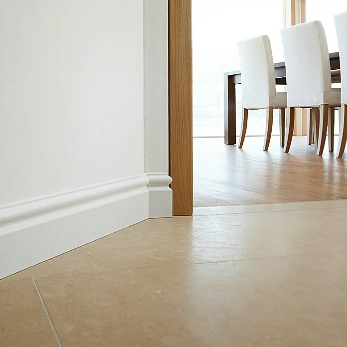 Sockelleiste FU116L - 240 cm x 15 mm x 115 mm, MDF, Weiß, Foliert - Detailaufnahme eines weißen Sockels an einer Wand, der einen eleganten Übergang zum Boden bildet. Im Hintergrund sind Stühle zu sehen.