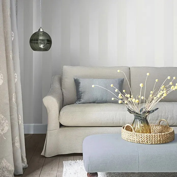 Perlmuttstreifen -  - Ein gemütliches Wohnzimmer mit hellbeigem Sofa, blauem Hocker und gestreifter Wand, akzentuiert durch eine graue Pendelleuchte.
