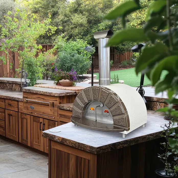 Domofire Kaminofen Capri Top - Material Verkleidung: Keramik, Weiß - Dekorativer Pizzaofen in Bronze und Creme mit Edelstahlschornstein auf einer Steinarbeitsplatte in einer Außenküche.