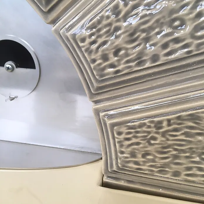Domofire Kaminofen Capri - Material Verkleidung: Keramik, Weiß - Gerätedetail: glänzendes, strukturiertes beiges Keramikteil neben einer silbernen Metallplatte mit Schraube.