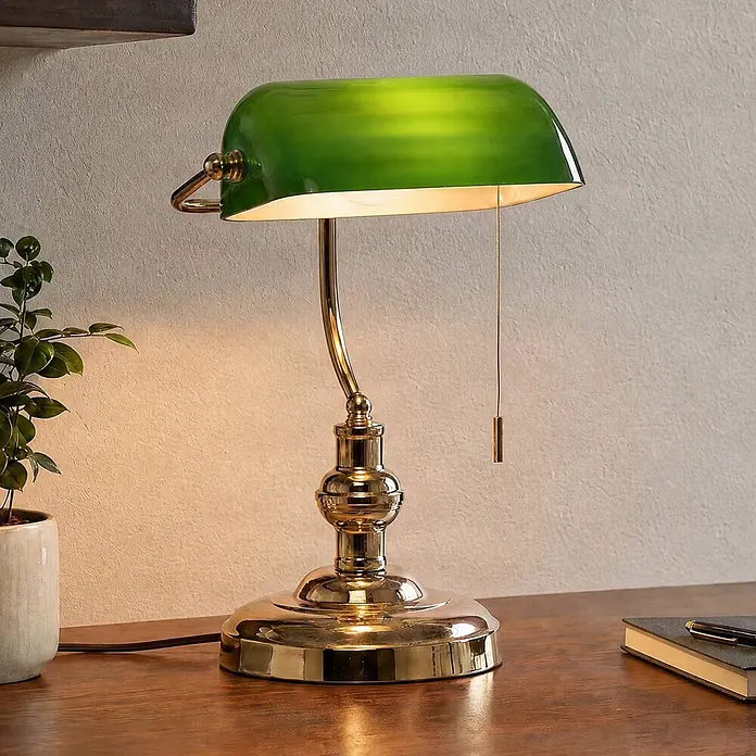 Lindby Tischleuchte Milenka - messing / gold, grün, E27 - Grün beschirmte Messing-Bankerlampe auf einem Holzschreibtisch, eingeschaltet und warmes Licht spendend.