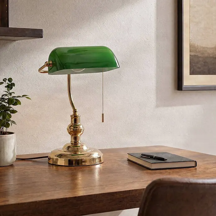 Lindby Tischleuchte Milenka - messing / gold, grün, E27 - Grüne Glas-Bankerlampe mit poliertem Messingfuß auf einem Holzschreibtisch mit Notizbuch.