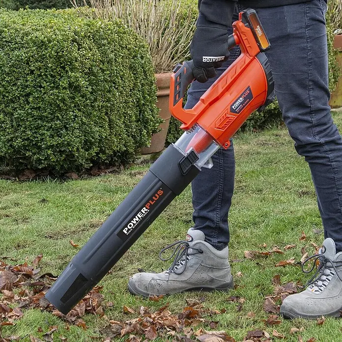 PowerPlus Soplador de batería POWDPGLBS1 - 20 V, 1 batería, 2 Ah, Velocidad de soplado: 145 km/h - Una persona usa un soplador de hojas inalámbrico naranja y negro para limpiar hojas de la hierba en un jardín.