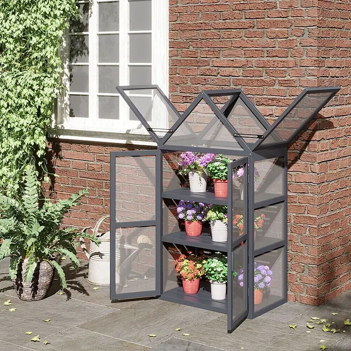 SONNENH Gewächshaus - 70,5 x 42 cm, Material Seitenwand: Kunststoff, Grau - Kleines graues Metall- und Klarpaneel-Gewächshaus mit mehreren Regalen voller bunter Topfpflanzen, im Freien an einer Ziegelwand.