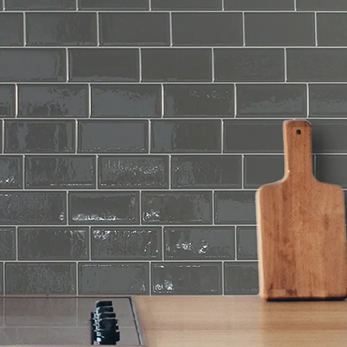 Smart Tiles Selbstklebemosaik Metro - 29,34 x 21,28 cm, Grigio, Glänzend - Glänzende dunkelgraue Keramik-Subway-Fliesen als Küchenrückwand, mit einem Holzschneidebrett davor.