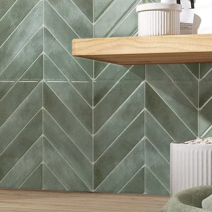 Smart Tiles Selbstklebemosaik Chevron - 29,02 x 22,86 cm, Solna, Glänzend - Grüne Keramik-Wandfliesen im Fischgrätmuster mit leichter Textur, hinter einem Holzregal und einer Arbeitsplatte.