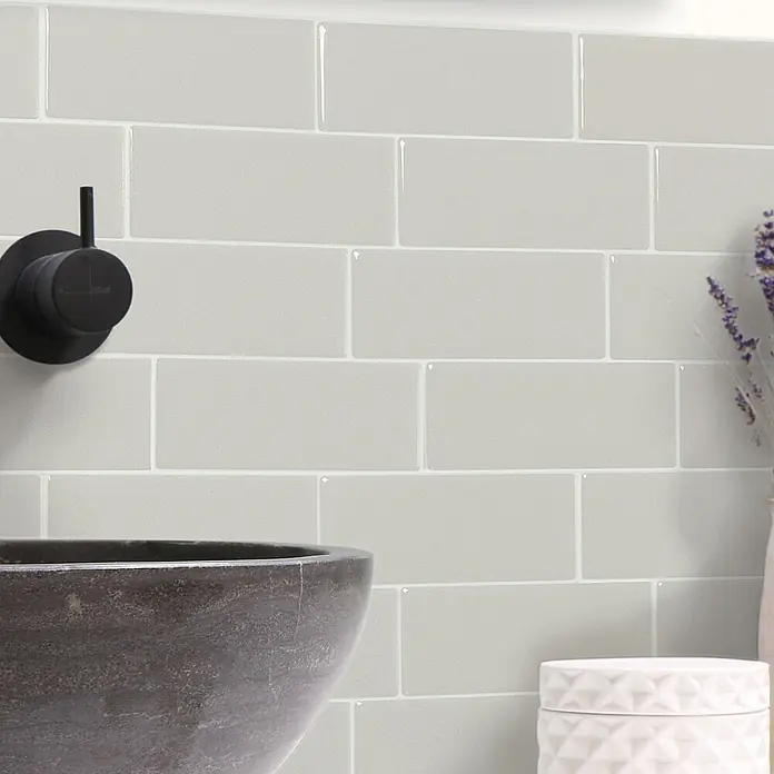 Smart Tiles Selbstklebemosaik Metro - 29,34 x 21,28 cm, Cassandra, Glänzend - Glänzende hellgraue Metro-Fliesen als Badezimmerwandverkleidung hinter dunklem Waschbecken und schwarzem Wasserhahn.