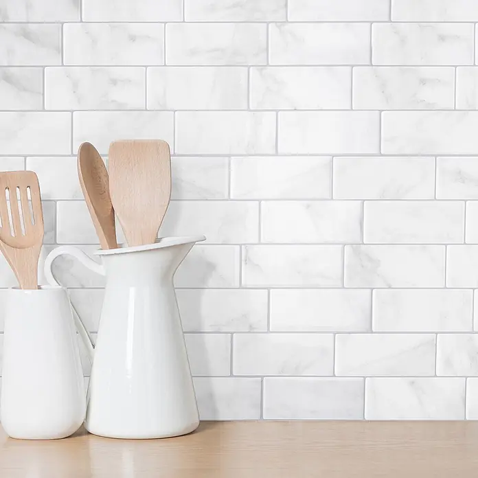 Smart Tiles Selbstklebemosaik Metro - 29,34 x 21,28 cm, Carrera, Glänzend - Weißer U-Bahn-Fliesen-Spritzschutz in Marmoroptik hinter Küchenutensilien aus Holz und weißen Behältern auf einer Arbeitsplatte.