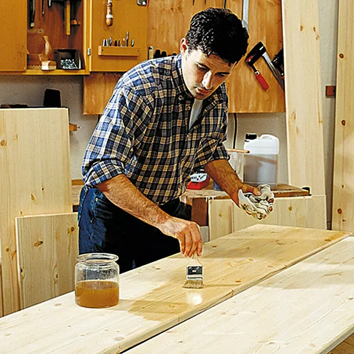 Do it wood Tablero de madera laminada - Pino, B/B, 200 x 25 x 1,8 cm - Un hombre en un taller está aplicando tinte para madera a una tabla de madera con un pincel.