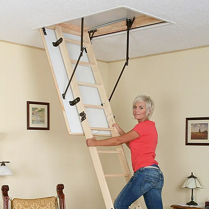 Star Stairs Bodentreppe Star Comfort - 120 x 60 cm, Wärmedämmung: 1,22 W/(m²K) - Eine Frau steigt eine hölzerne Dachbodentreppe hinauf, die aus einer Deckenöffnung ausfährt. Die Treppe ist mit einem weißen Paneel und schwarzen Beschlägen versehen.