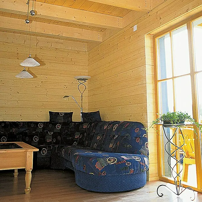 Friso machihembrado - 2.700 x 121 x 14 mm - Sala de estar con sofá seccional, paredes de madera y ventanas. El sofá es azul estampado, la habitación es luminosa y acogedora.