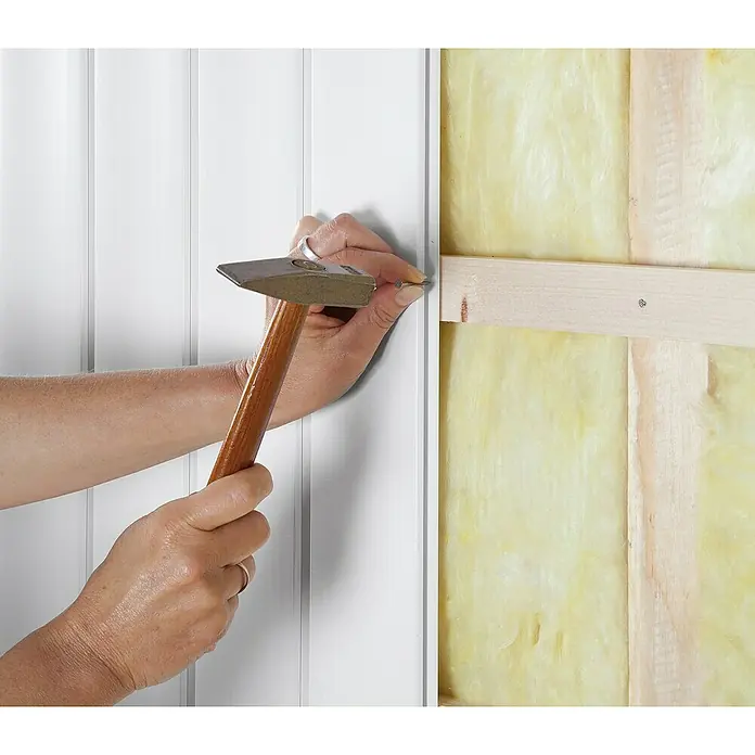 BaukulitVox Basic-Line Wandpaneel B25 - Weiß, 3.000 x 255 x 10 mm - Ein Handwerker befestigt mit einem Hammer eine weiße Holzvertäfelung an einer Wand mit gelber Dämmung. Werkzeuge und Renovierung im Fokus.