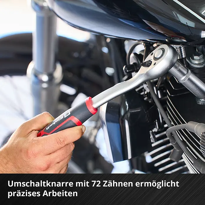 Einhell Werkzeugkoffer E-Case KFZ - 121 -tlg. - Hand benutzt eine silberne Umschaltknarre mit schwarz-rotem Griff an einem Motorradmotor. Die 72-Zahn-Umschaltknarre ermöglicht präzises Arbeiten.