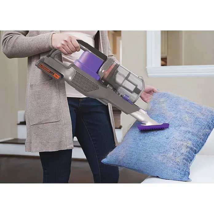 Black+Decker Aspirador de batería Power Series Extreme - 18 V, 2 Ah, Volumen de recipiente: 650 ml - Persona usa una aspiradora de mano inalámbrica gris y morada con accesorio para pelo de mascota en un cojín azul.