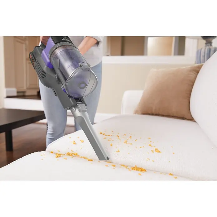 Black+Decker Aspirador de batería Power Series Extreme - 18 V, 2 Ah, Volumen de recipiente: 650 ml - Persona usa una aspiradora de mano gris y morada con depósito transparente para limpiar migas naranjas de un sofá blanco.