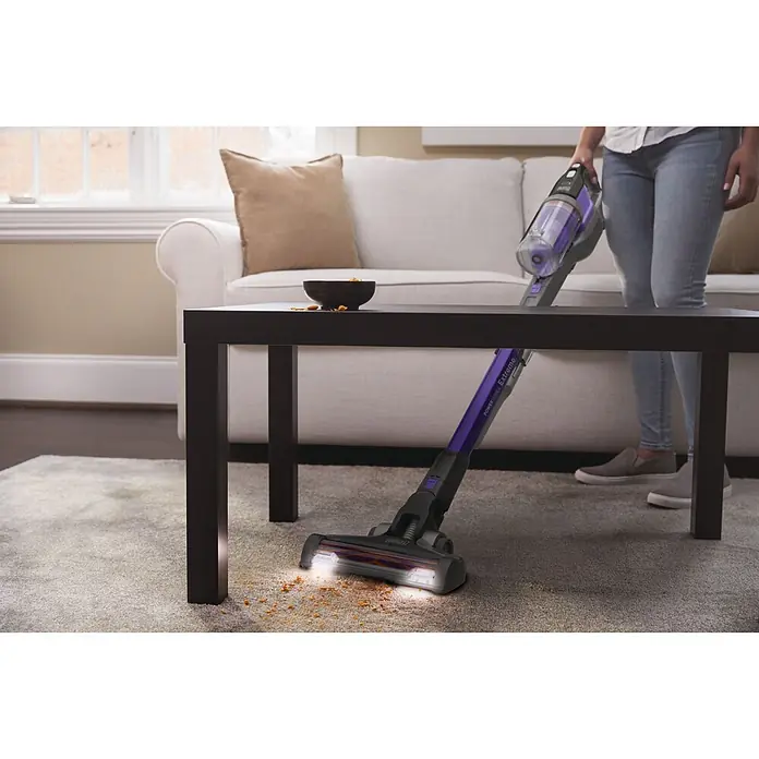 Black+Decker Aspirador de batería Power Series Extreme - 18 V, 2 Ah, Volumen de recipiente: 650 ml - Una persona usa una aspiradora de escoba morada y negra para limpiar comida derramada de una alfombra clara bajo una mesa oscura.
